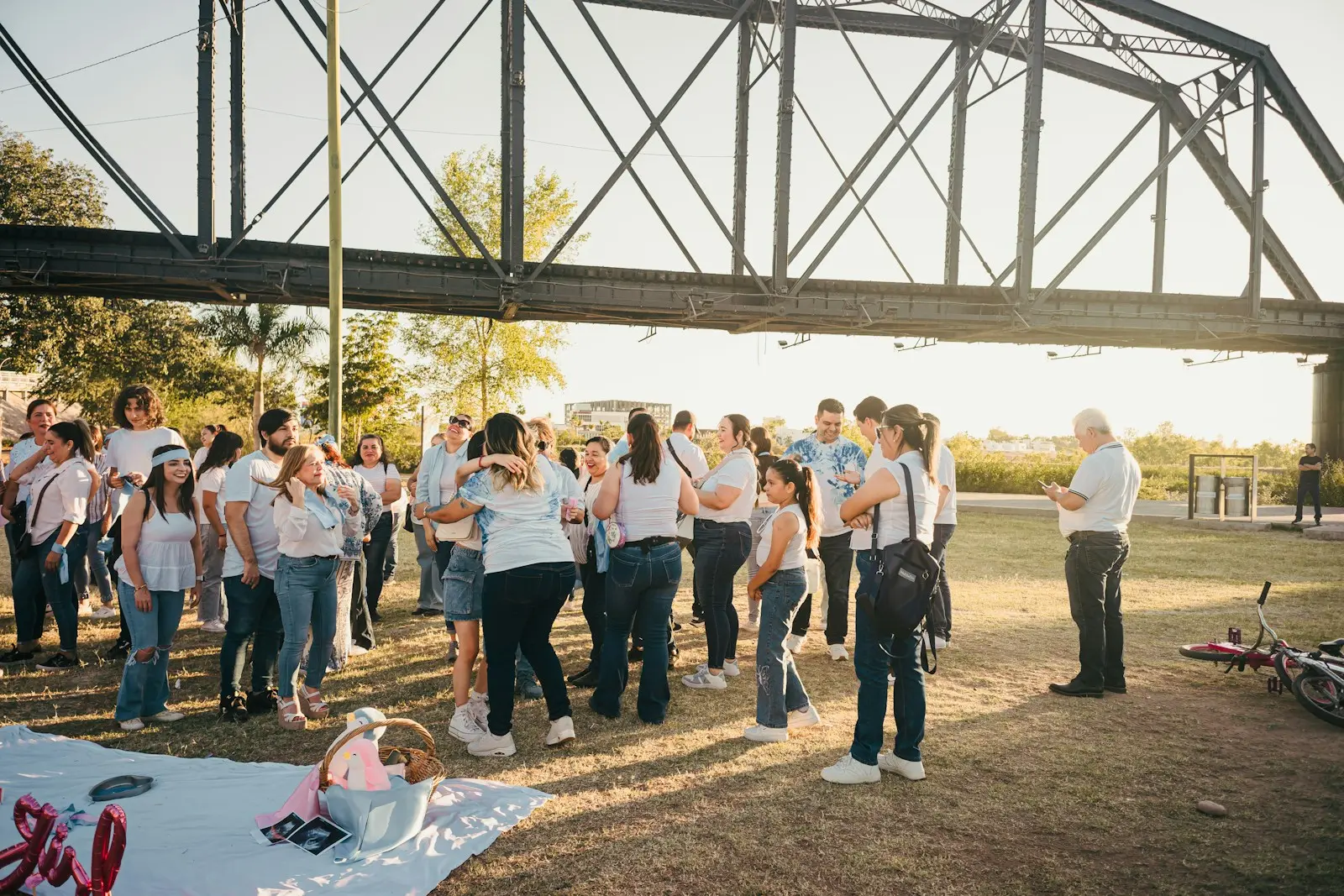 A diverse group of people dressed in white and light blue gather under a metal bridge, celebrating together with joy and hugs in a sunny park setting.