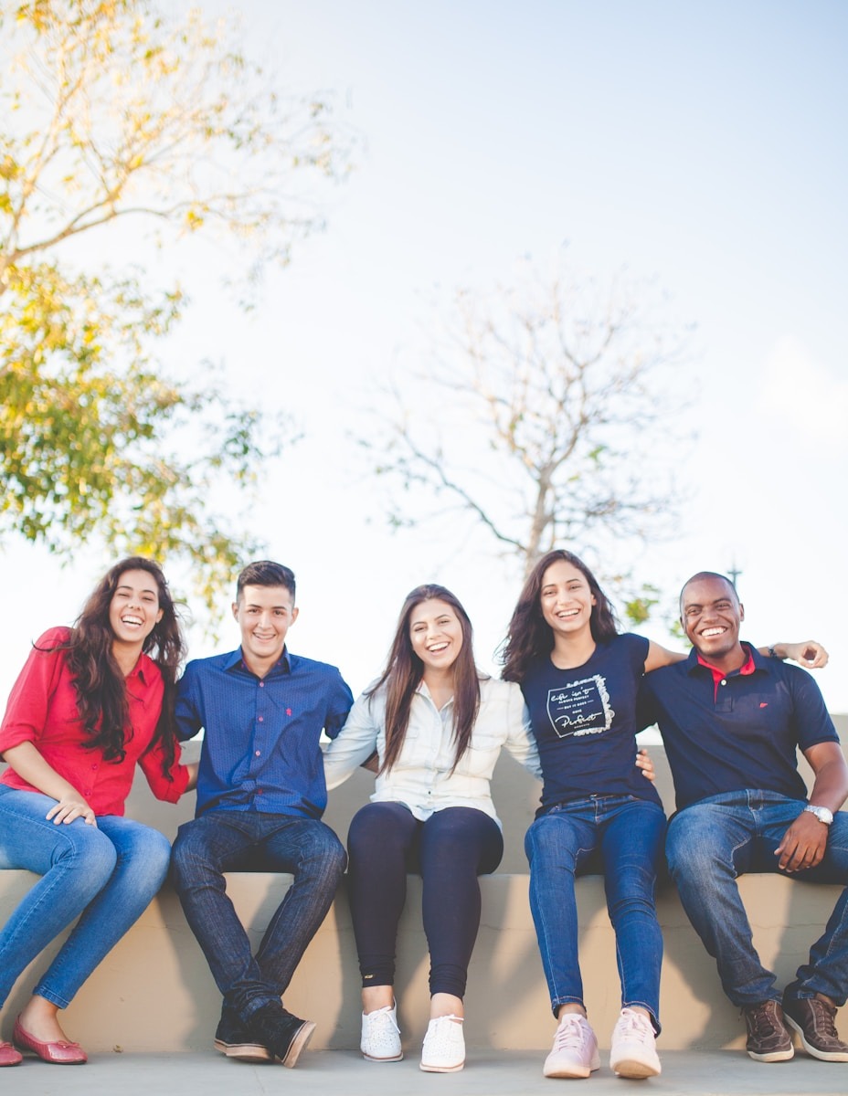A diverse group of five young adults smiles and sits together outdoors, showcasing friendship and joy in a lively, sunny setting.
