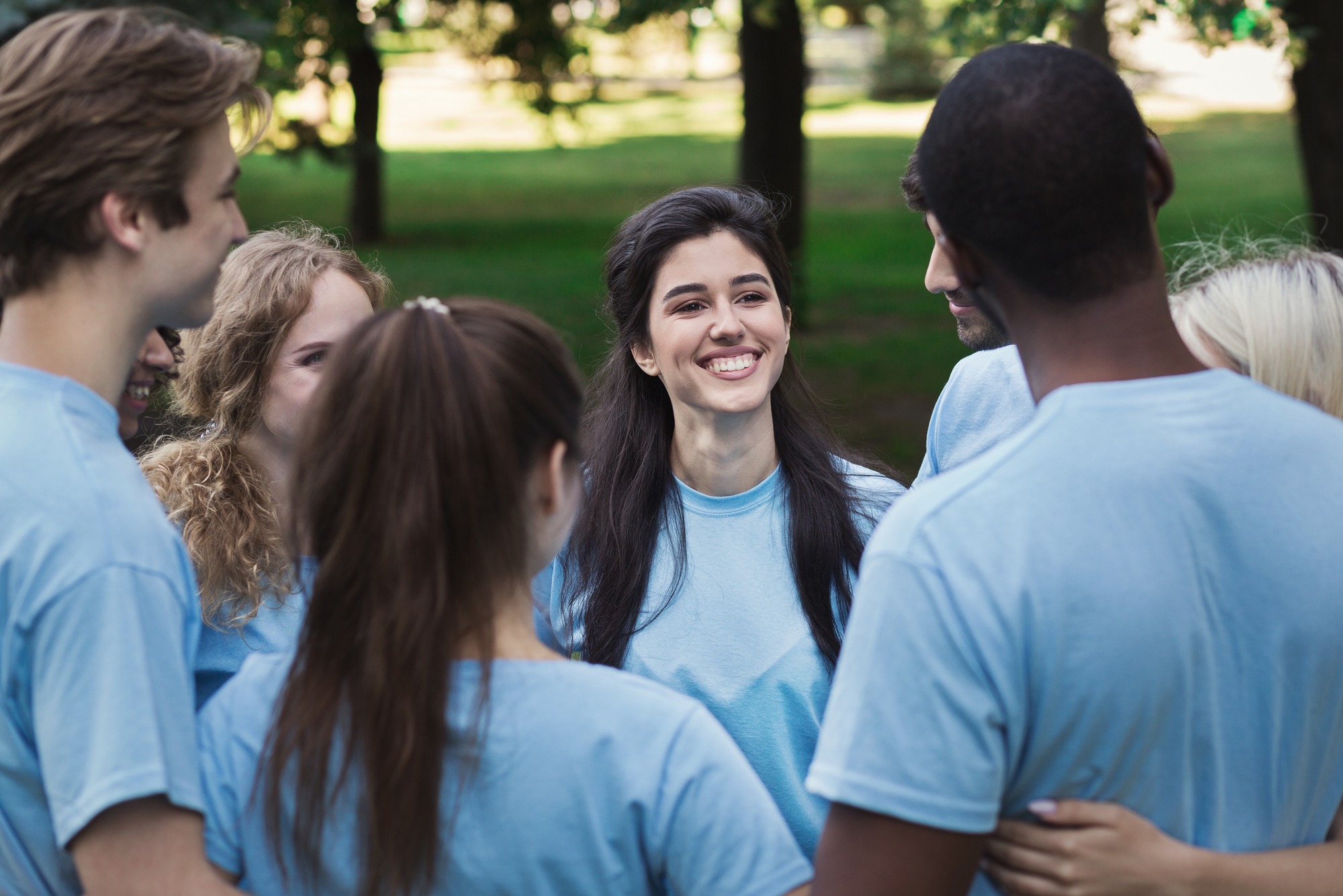 A diverse group of young adults in light blue shirts smiles and interacts in a park, showcasing camaraderie and teamwork.
