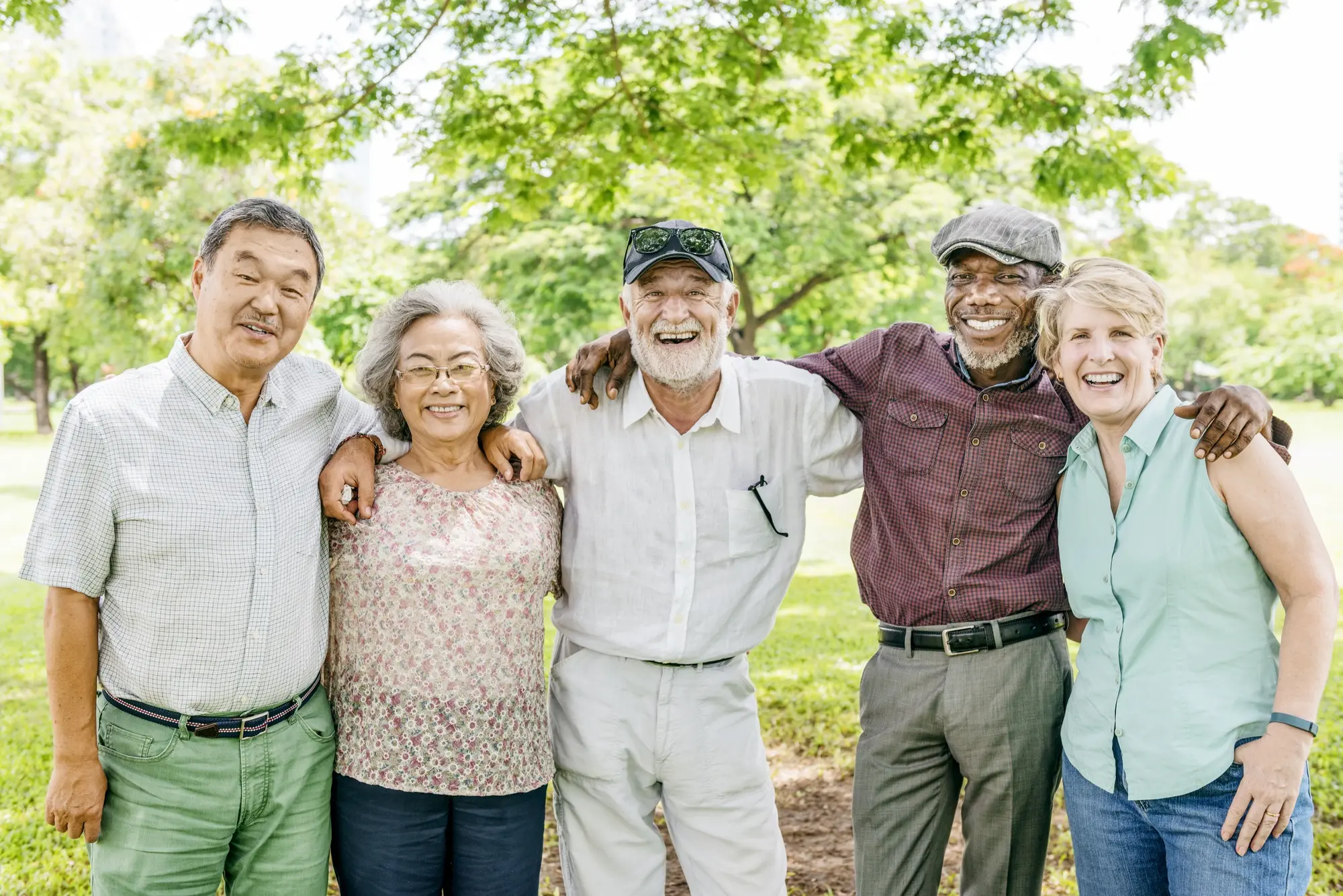 A diverse group of five seniors smiles together in a park, showcasing friendship and joy in a sunny outdoor setting.